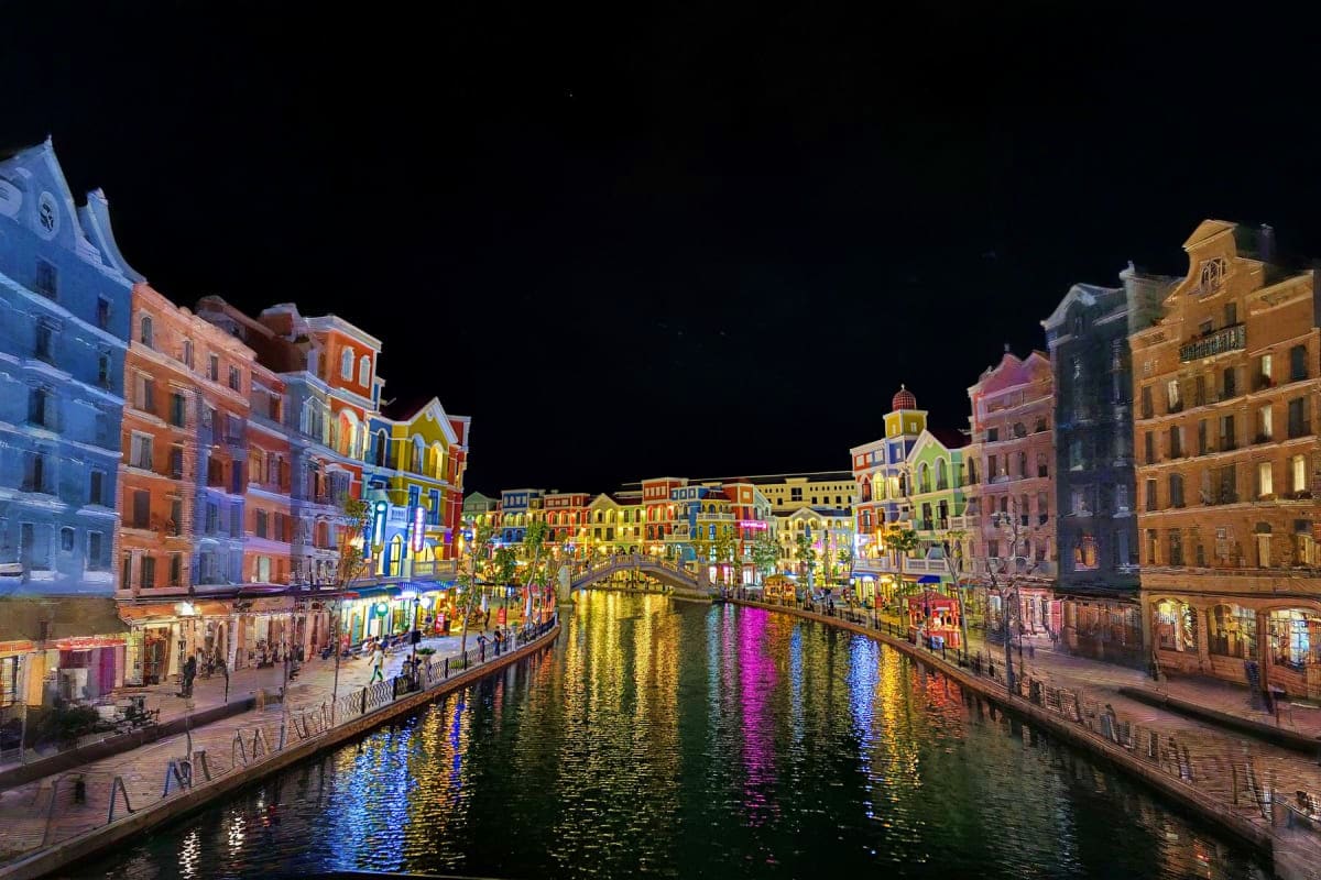 Halong Waves and Phu Quoc Days Illuminated waterfront buildings reflected in a canal at night with colorful lights and pedestrians along the promenade