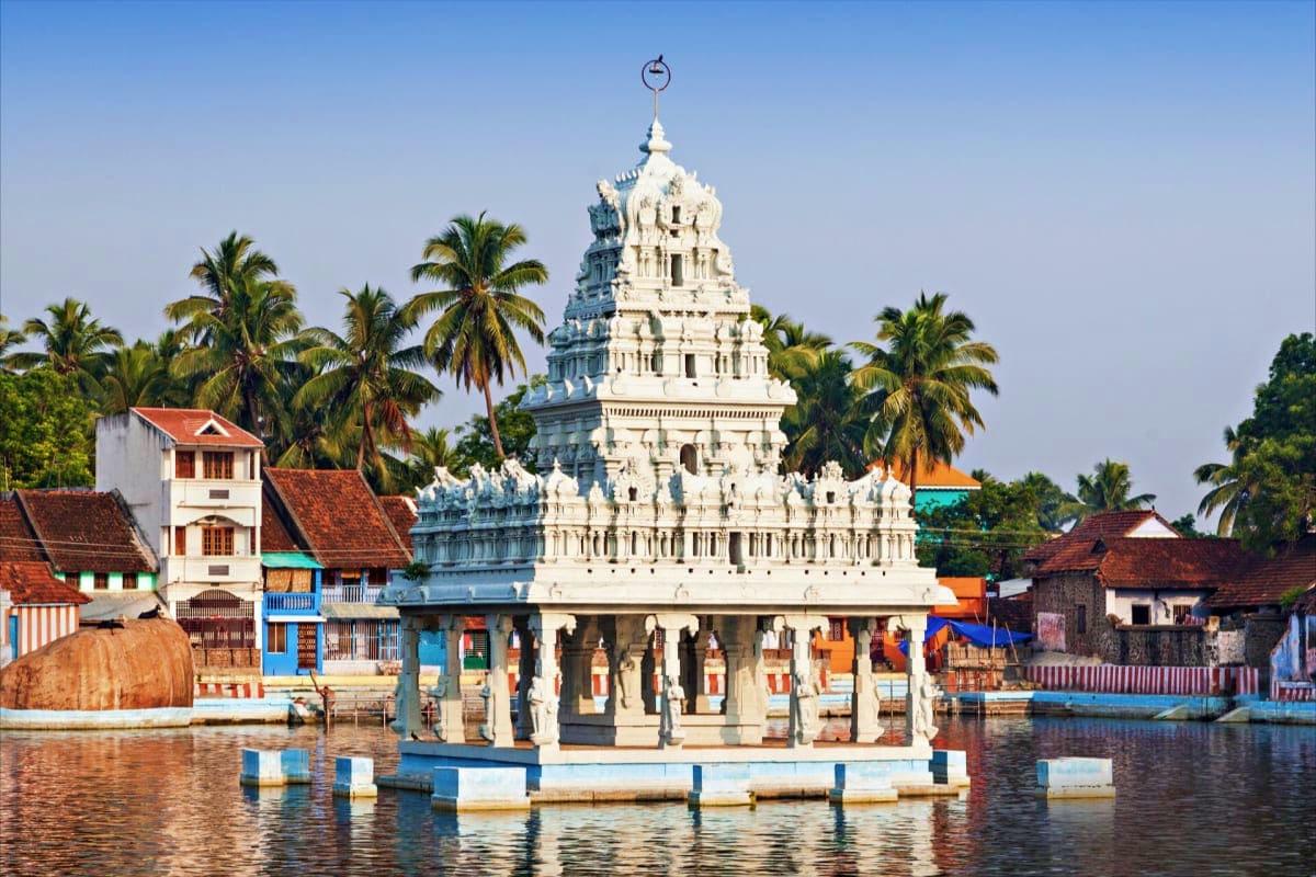 White Hindu temple with ornate tower reflected in Kerala backwater, surrounded by palm trees and traditional buildings