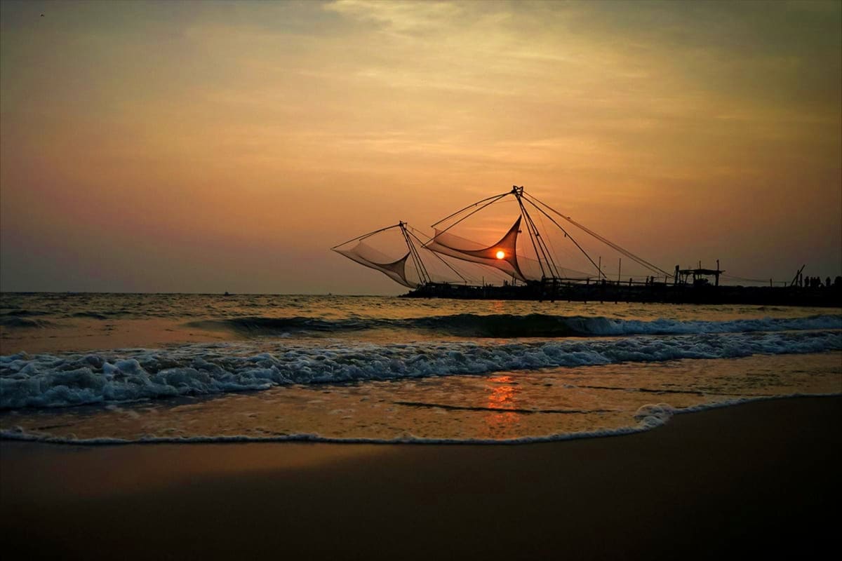 Traditional Chinese fishing net at sunset on a Kerala beach with waves and golden sky