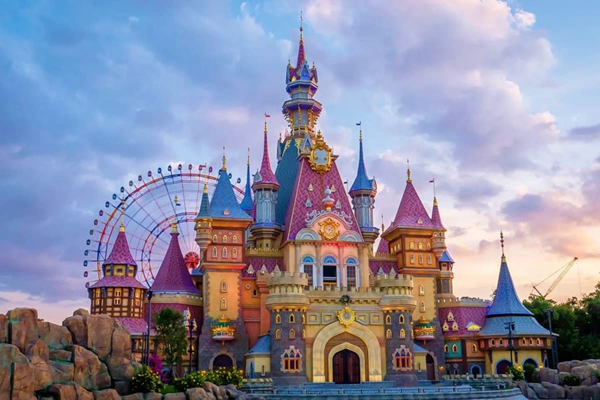 Colorful fairy tale castle with purple spires, golden accents, and Ferris wheel at sunset in theme park setting