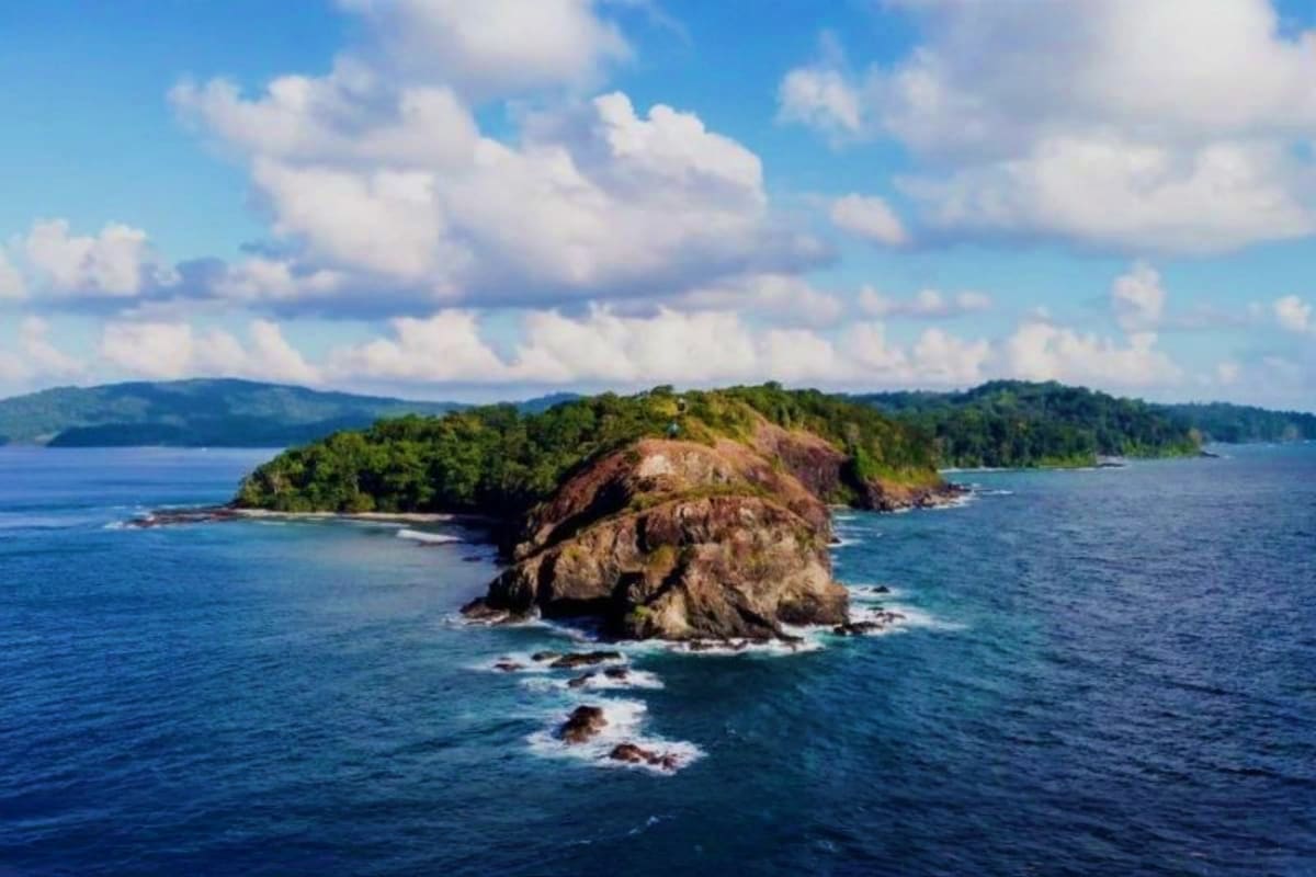 Rocky forested island with steep cliffs surrounded by blue ocean water and distant coastline under cloudy sky