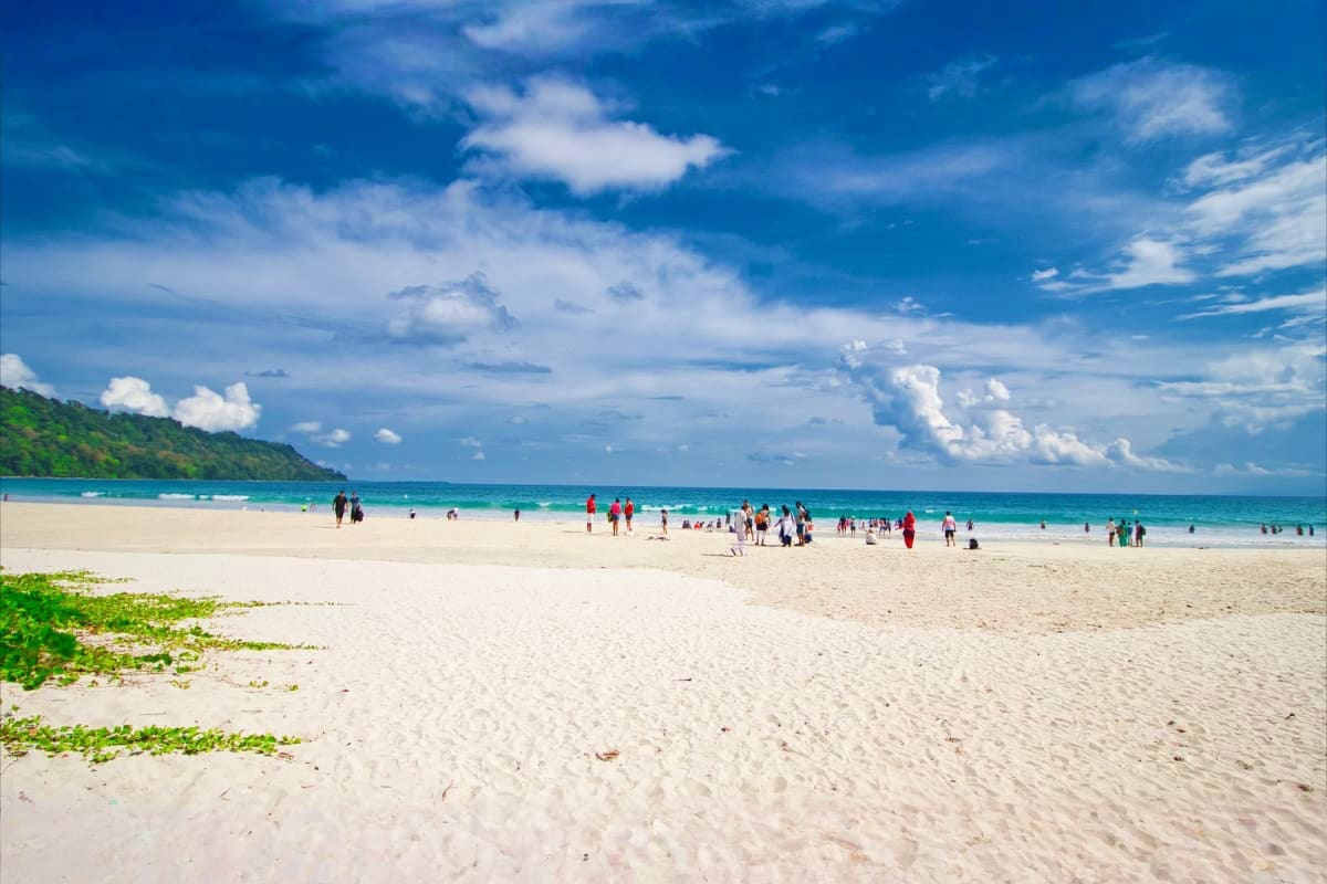 Quick Andaman Escape History Beaches and Island Tours Tropical beach in Andaman with white sand, turquoise waters, visitors swimming and relaxing, forested hillside, blue sky with