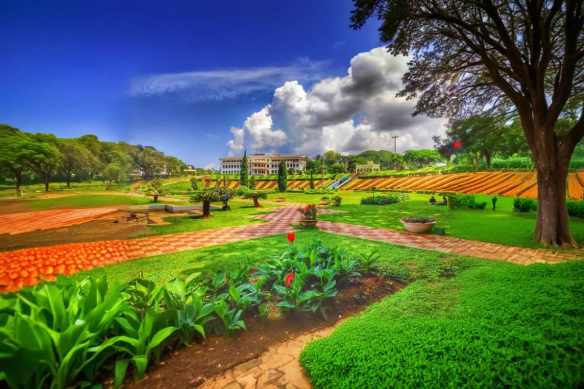 Lush garden with red tulips, manicured hedges, brick pathways, and colonial-style palace buildings under blue sky with white 