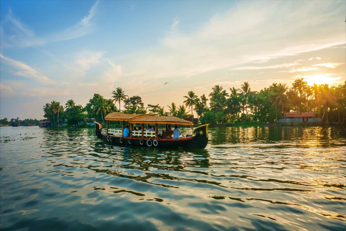 Traditional Kerala houseboat with golden roof sailing on backwaters at sunset, surrounded by palm trees and calm water