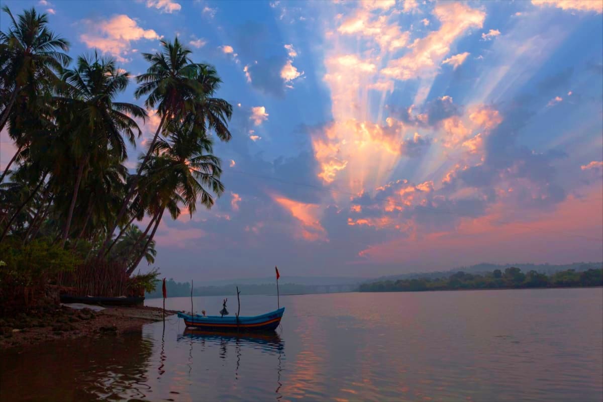 Blue and orange fishing boat on calm water at sunset with palm trees and dramatic sky with golden clouds reflecting on water 
