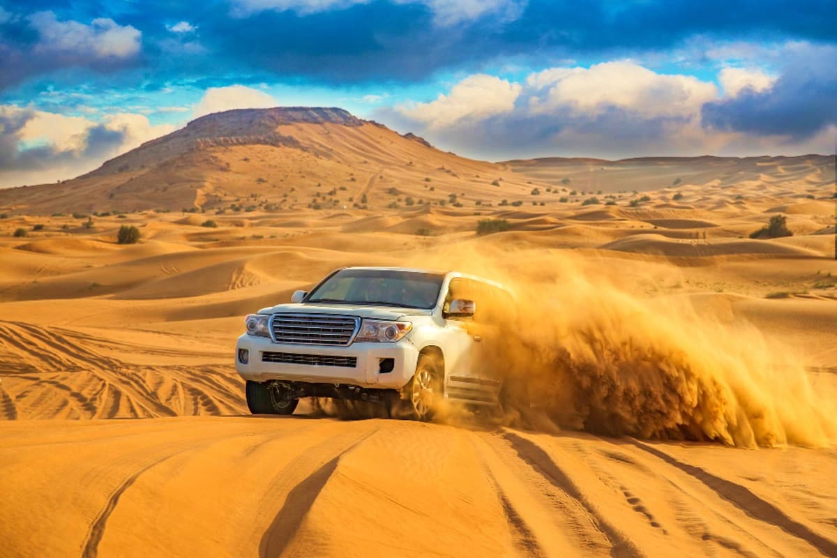 White SUV driving through sand dunes with dust cloud, desert mountain landscape under blue sky
