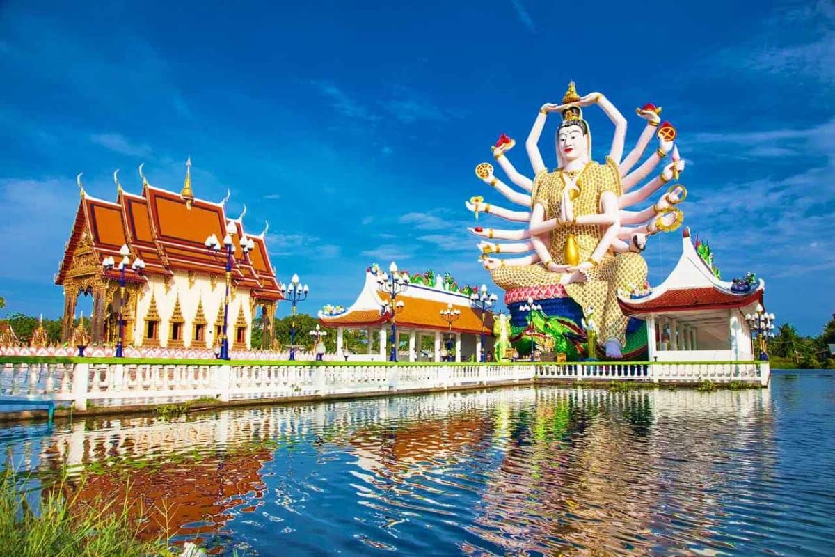 Exclusive Koh Samui Thai Buddhist temple with multi-armed golden deity statue beside ornate red-roofed buildings reflected in water under blue sk