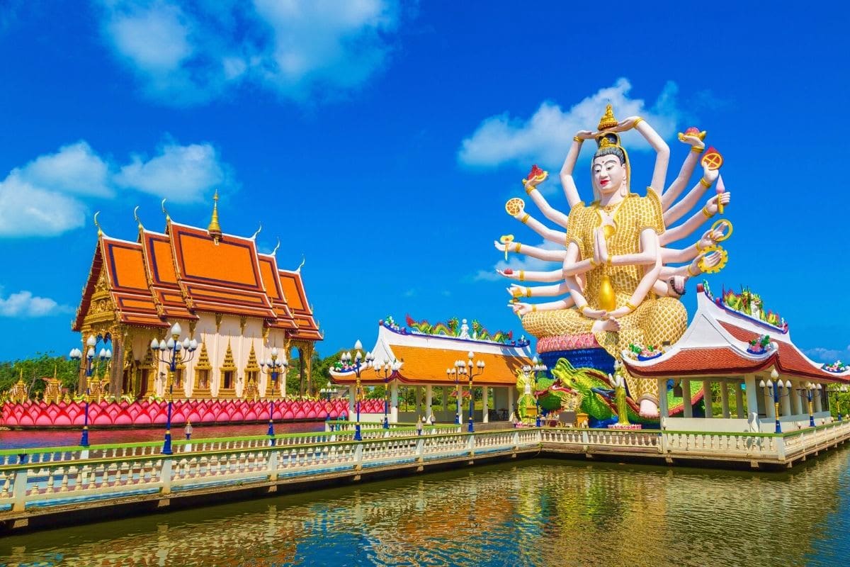 Exclusive Koh Samui Thai Buddhist temple with golden multi-armed deity statue beside ornate orange-roofed shrine reflected in canal water under b