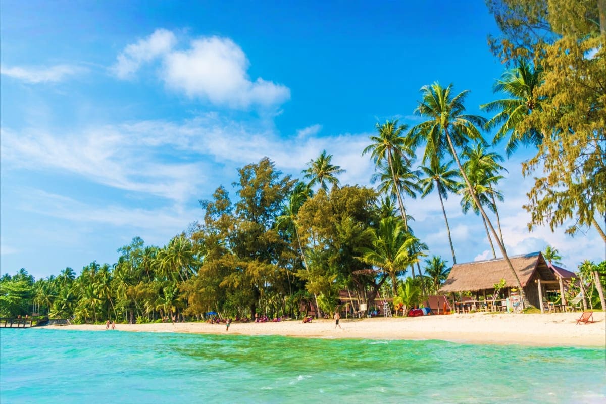 Tropical Port Blair Getaway 4 Days Island Experience Tropical beach in Port Blair with turquoise water, white sand, palm trees, and traditional thatched-roof structures along the