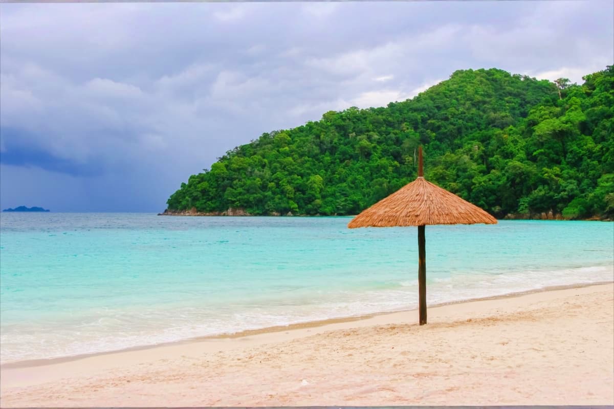 Tropical Port Blair Getaway 4 Days Island Experience Thatched umbrella on pristine sandy beach with turquoise water and dense green forested island in background