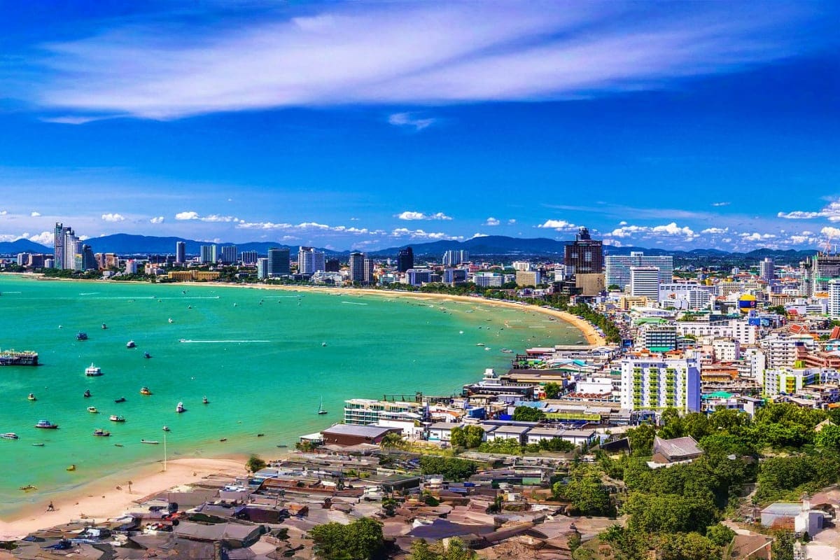 Aerial view of Pattaya beach with turquoise waters, sandy shore, moored boats, and cityscape with mountains under blue sky