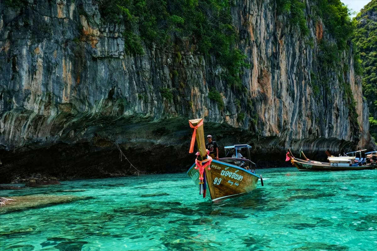 Traditional longtail boat with red and yellow fabric in turquoise water beneath towering limestone cliffs in Thailand