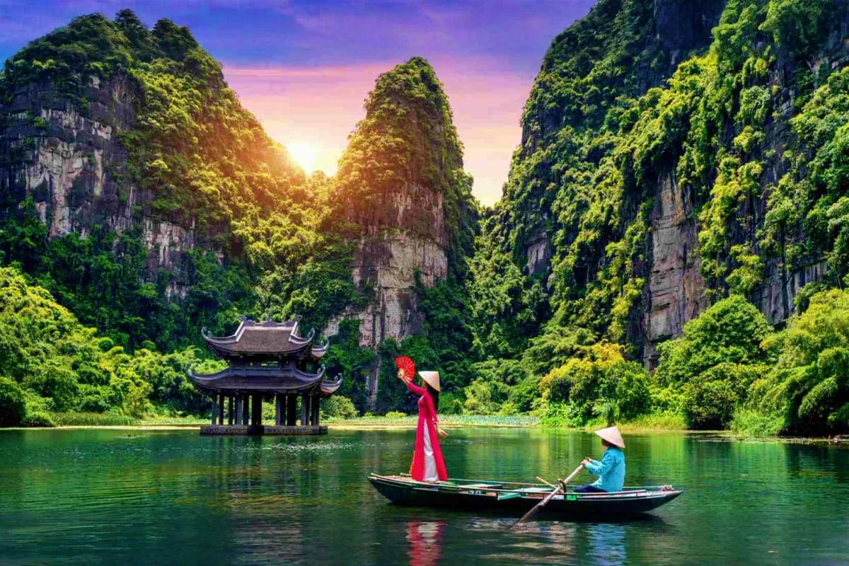 Woman in red áo dài on wooden boat with boatman in traditional cone hat, floating past limestone cliffs and pagoda at sunset 