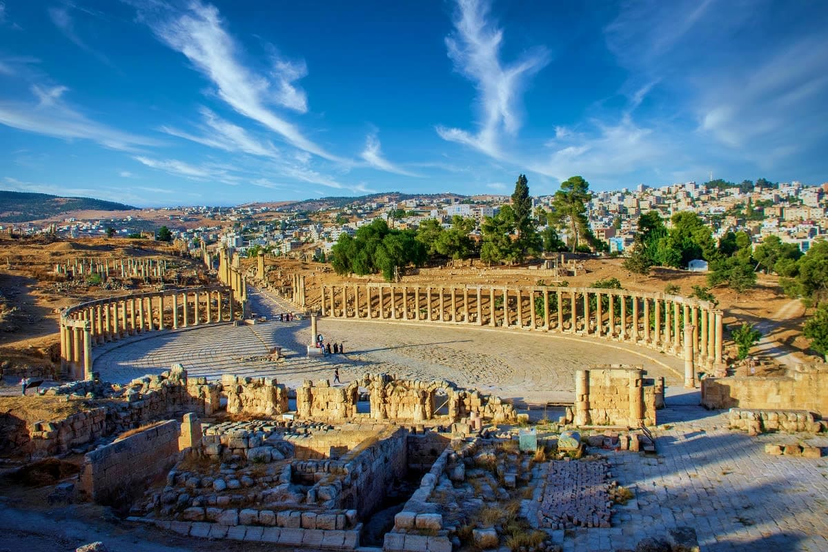 Ancient Roman amphitheater with columned seating surrounded by stone ruins, overlooking hillside city with modern buildings u