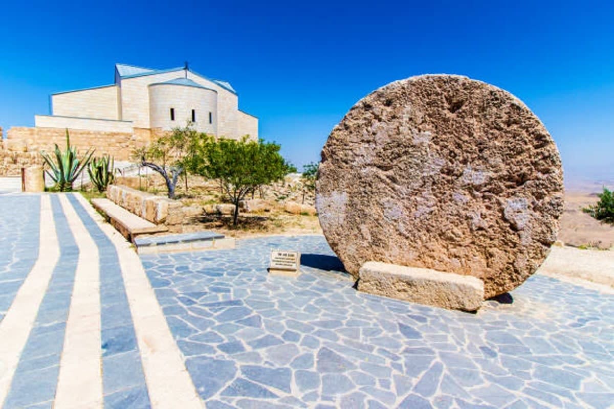 Ancient stone wheel monument with white church building and paved courtyard under clear blue sky in Jordan