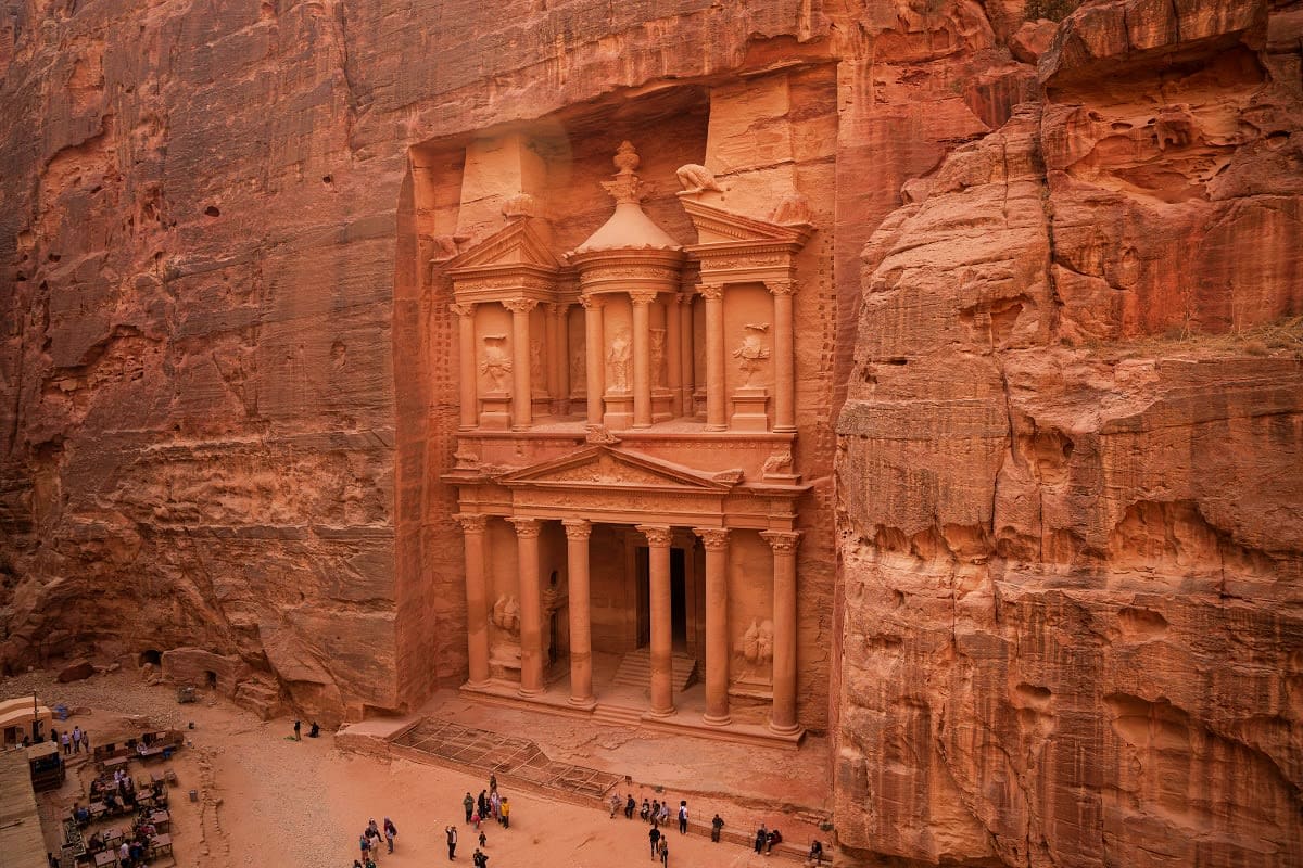 The Treasury, an ornate Nabatean temple facade carved into rose-red sandstone cliffs at Petra, Jordan, with tourists explorin