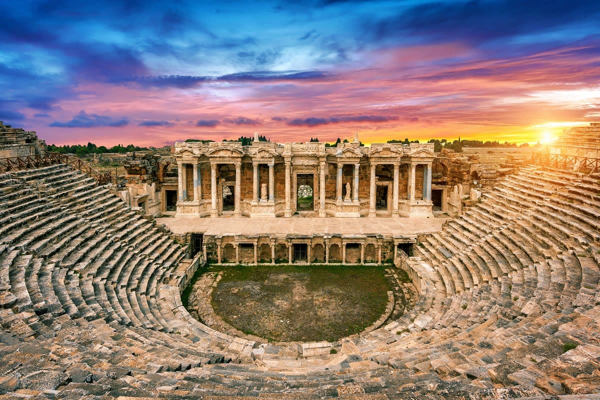 Ancient Roman amphitheater at sunset in Jerash, Jordan, with tiered stone seating and columned stage building under golden sk