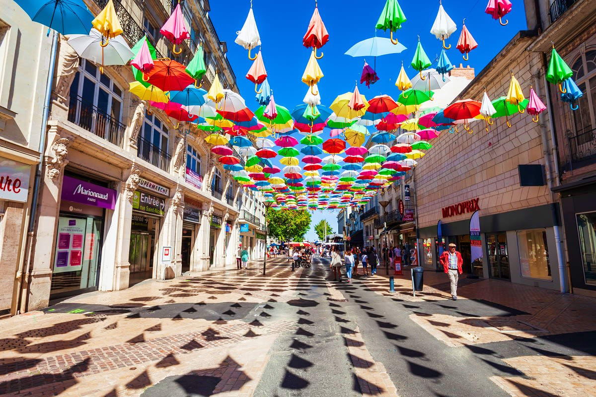 Colorful umbrellas suspended above a European shopping street with historic buildings and pedestrians on a sunny day