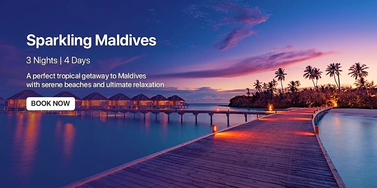 Twilight sunset over a Maldives resort with illuminated overwater bungalows, wooden boardwalk, and palm trees lining the trop