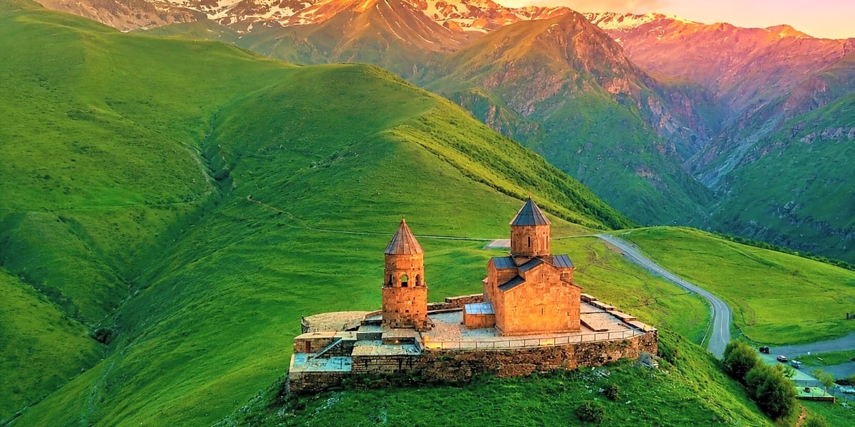 Tsminda Sameba Church with red stone towers set on green hillside surrounded by Caucasus Mountains at sunset in Georgia