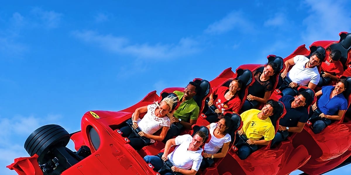 Group of diverse passengers enjoying a red roller coaster ride against a clear blue sky at an amusement park