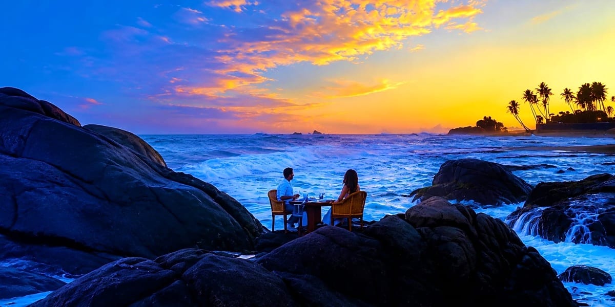 Couple dining at a table on rocky beach at sunset with ocean waves and palm trees in background