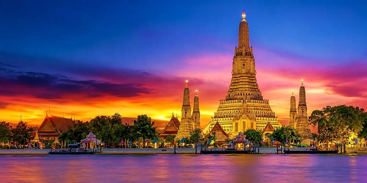 Wat Arun temple illuminated at sunset with golden spires reflected in the Chao Phraya River, Bangkok Thailand