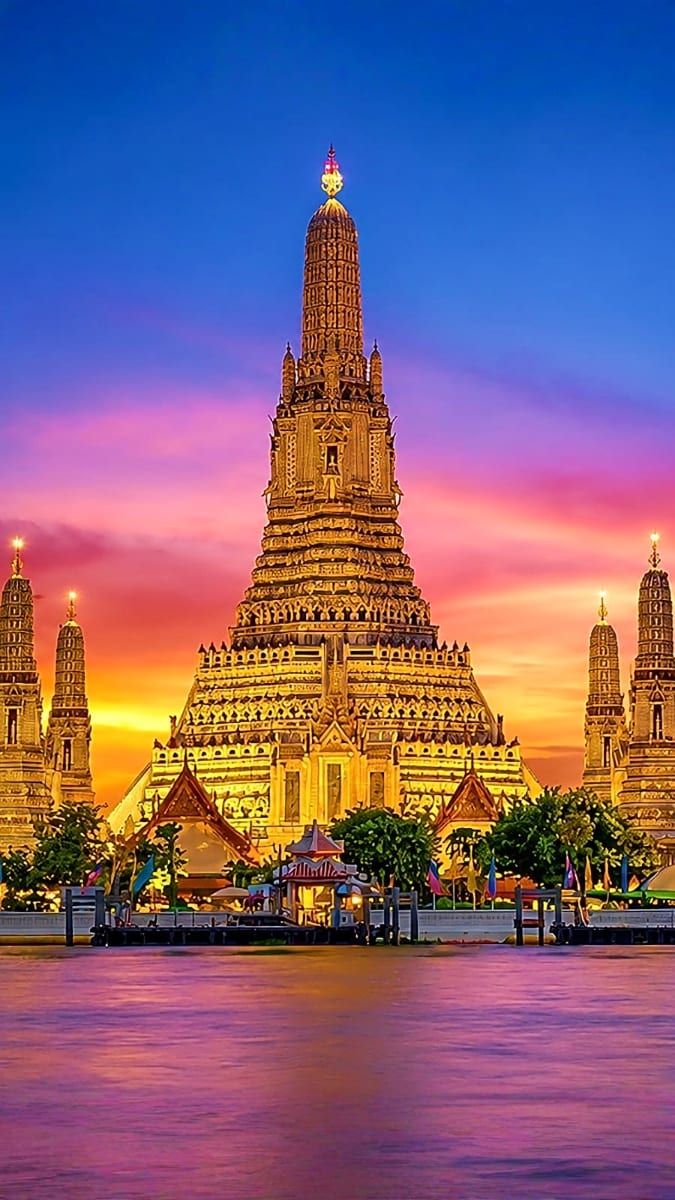 Wat Arun temple illuminated at sunset along the Chao Phraya River in Bangkok, Thailand, with golden spires reflected in the w