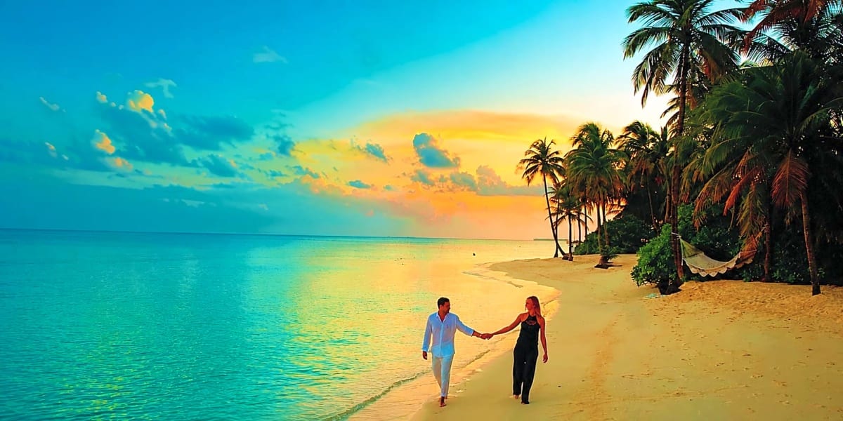 Couple holding hands walking on pristine sandy beach at sunset with palm trees and turquoise ocean water in Malaysia