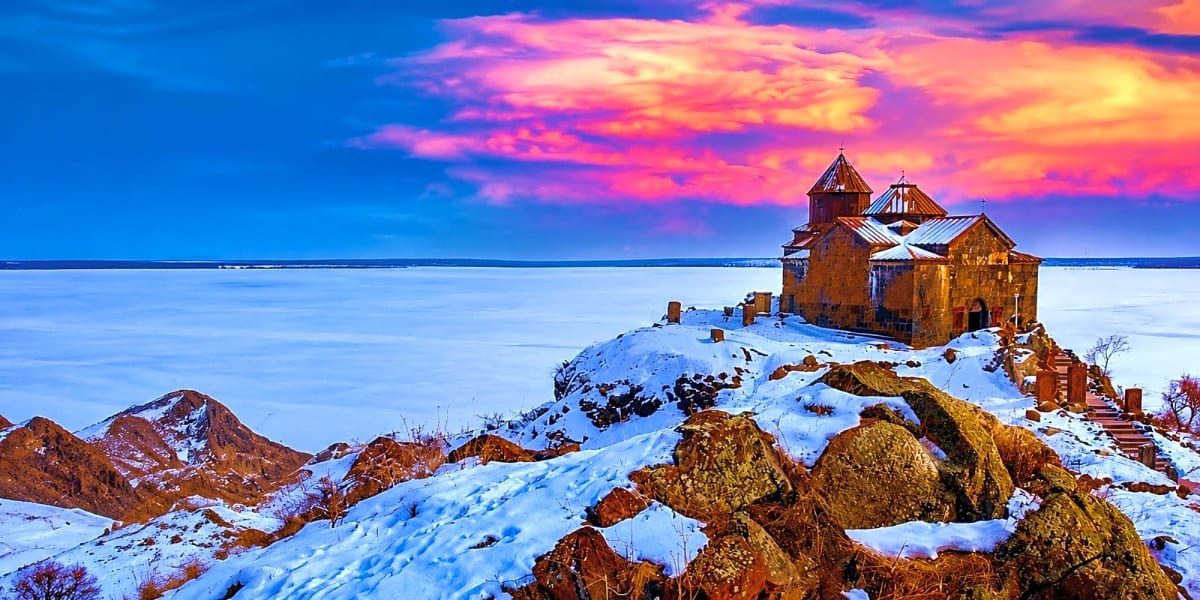 Armenian medieval monastery perched on snowy rocky hilltop overlooking frozen lake at sunset with vibrant pink and orange sky