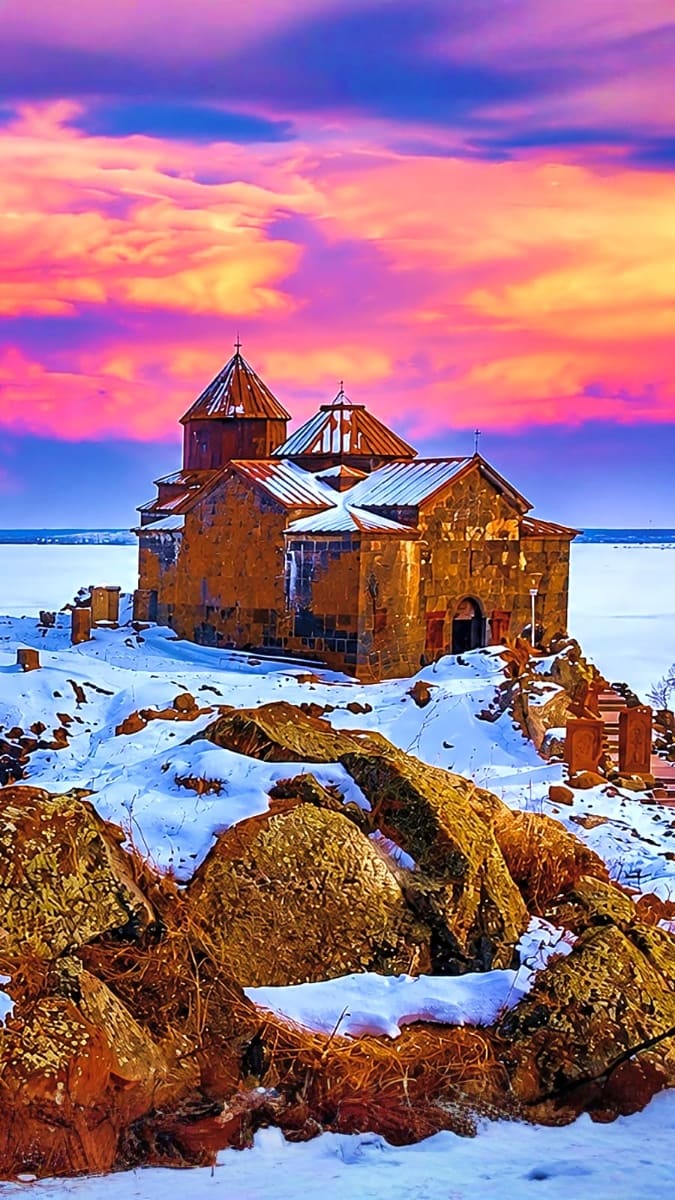 Ancient Armenian church with domed towers on rocky island surrounded by frozen lake at sunset with vibrant pink and purple sk
