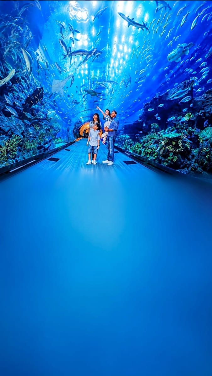 Family walking through underwater tunnel at Dubai Aquarium surrounded by sharks, rays, and tropical fish in blue ocean water