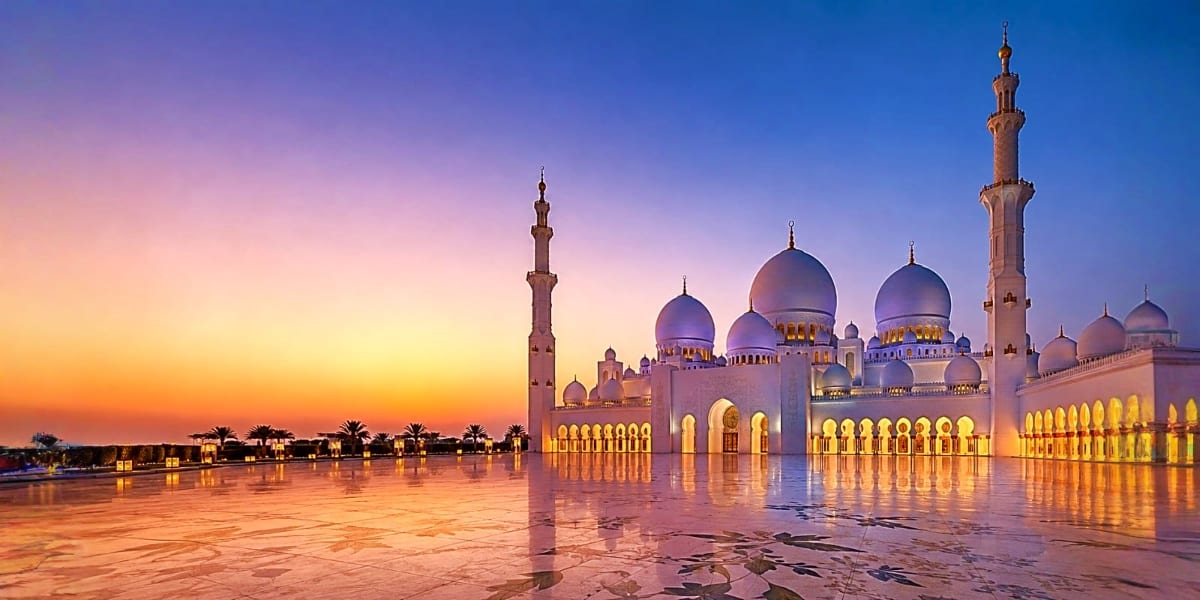 Sheikh Zayed Grand Mosque at sunset with illuminated white domes and minarets reflected in wet courtyard floor