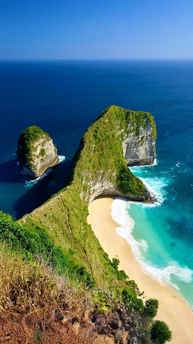 Aerial view of Kelingking Beach in Bali with turquoise waters, white sand beach, and distinctive green-topped cliff formation