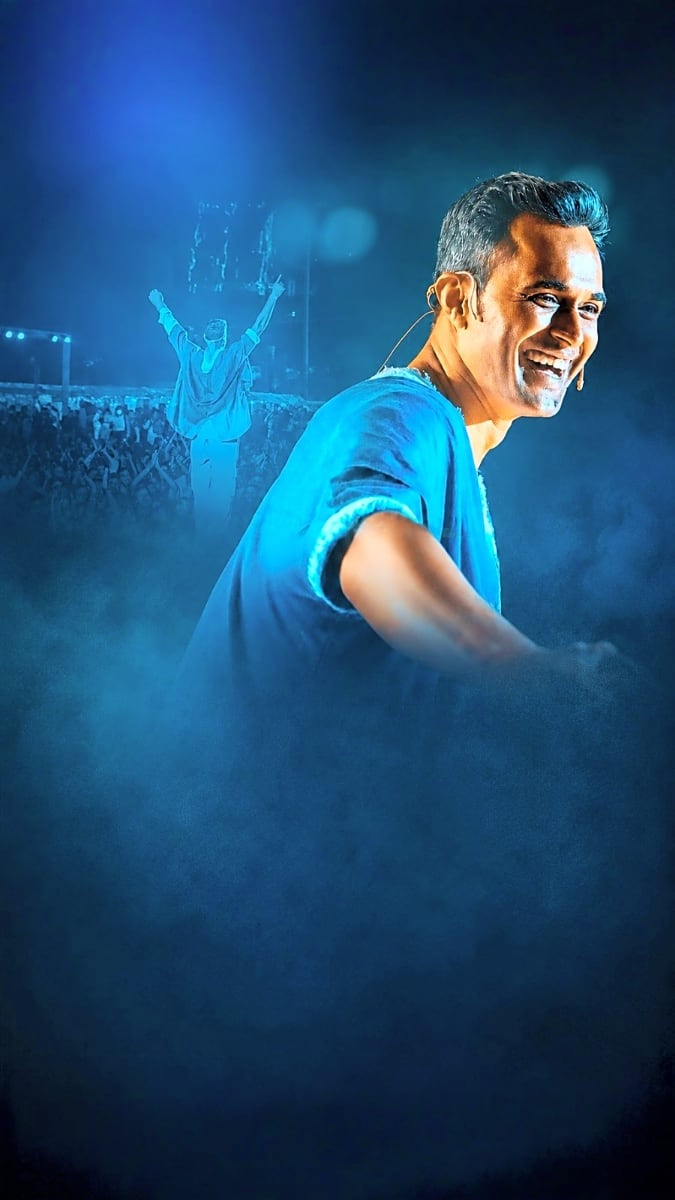 Male performer in blue shirt smiling at camera with concert crowd and stage backdrop in blue lighting