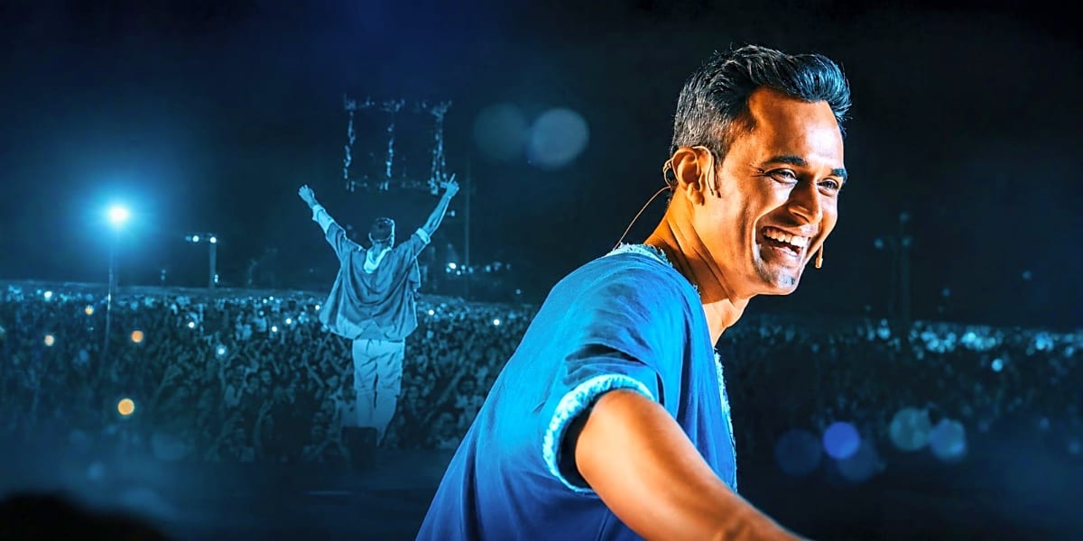 Man in blue shirt performing on stage at night with large crowd and illuminated historic clock tower in background