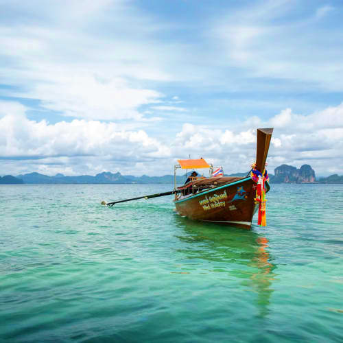 Traditional wooden long tail boat anchored in turquoise water with limestone cliffs and cloudy sky in Thailand