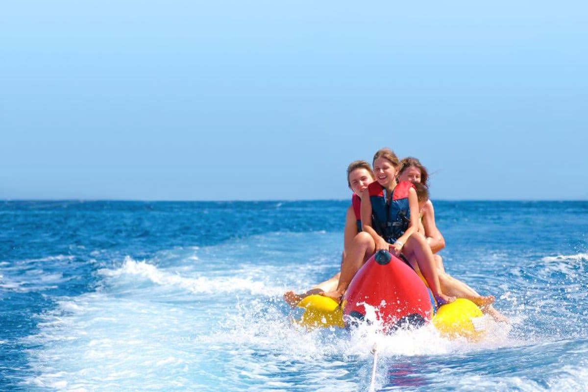 Banana Boat Ride in Dubai 15 Min Three children on a red banana boat with yellow handles riding through ocean waves under clear blue sky