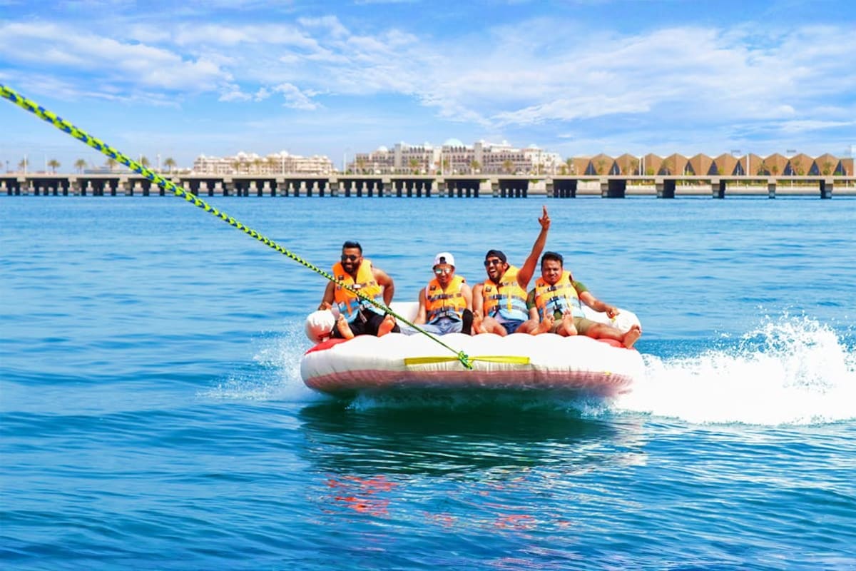 Donut Ride In Dubai 15 min Four people in yellow life jackets riding on a white inflatable donut tube being towed behind a boat in Dubai with the pier a