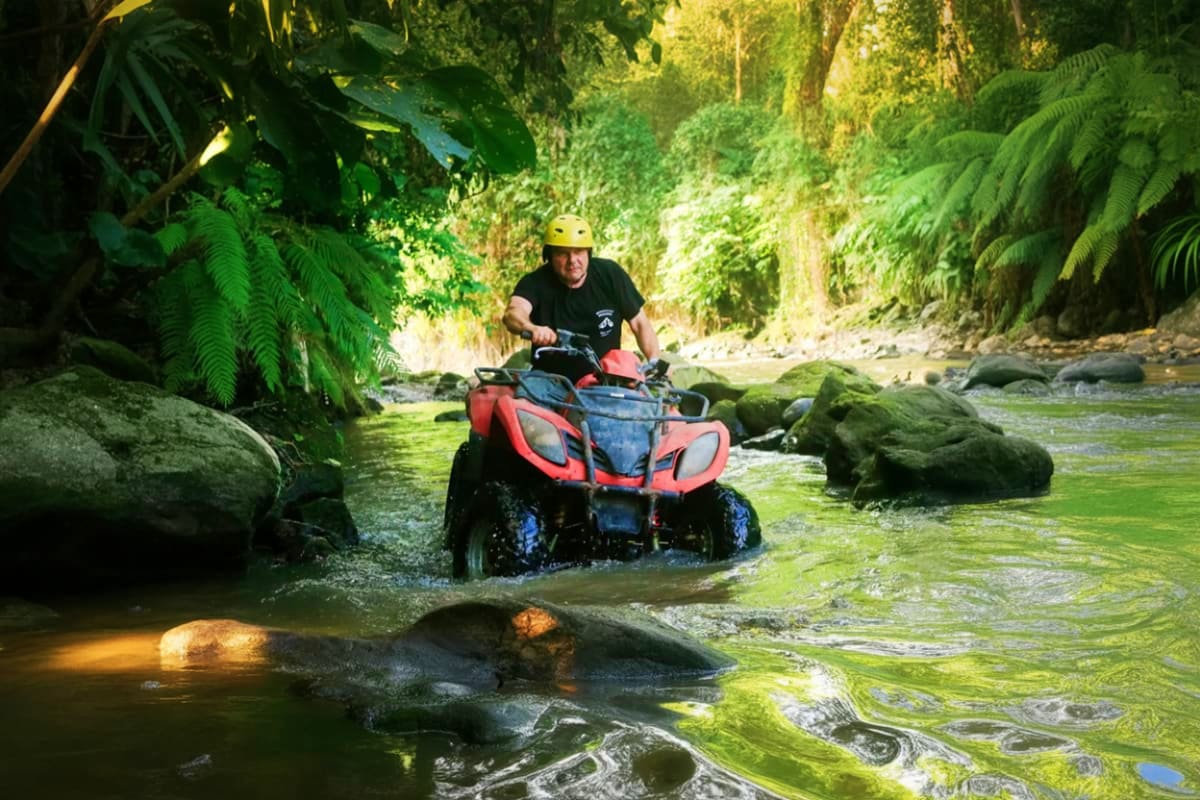 Man riding red ATV through shallow river surrounded by lush green jungle and ferns in Bali