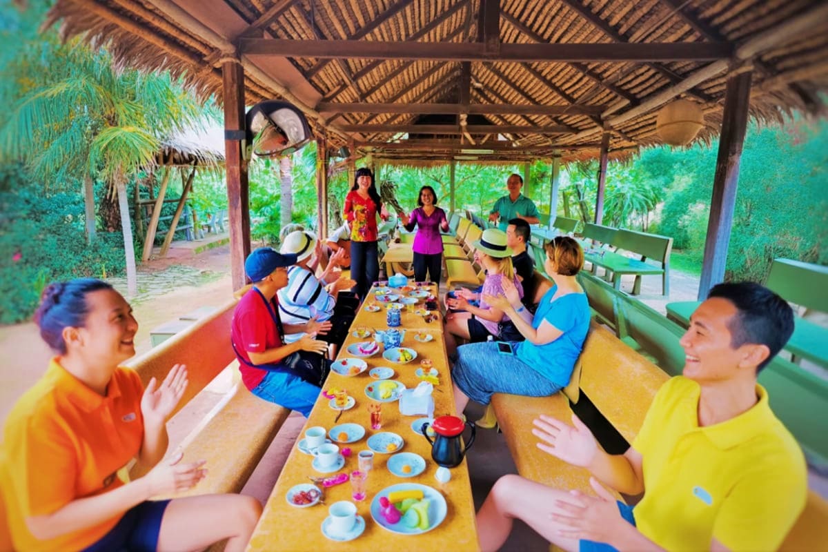 Mekong Delta My Tho City With Private Transfer Group of tourists dining together at long table under thatched roof structure in Mekong Delta rural setting with lush green s
