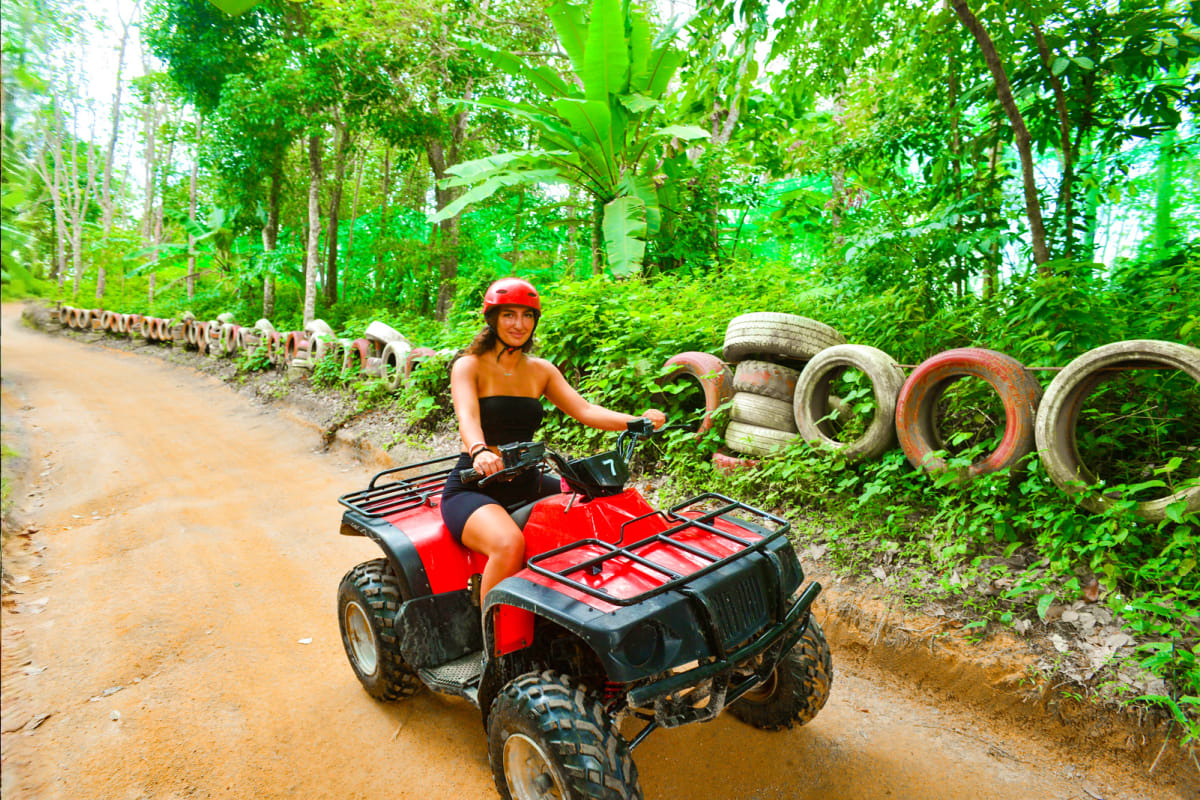 Woman riding red ATV on muddy jungle trail in Phuket with lush green forest and old tires on roadside