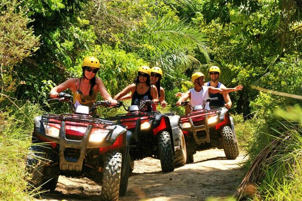 Group of five people riding red and black ATVs on a jungle trail through dense green vegetation wearing yellow safety helmets