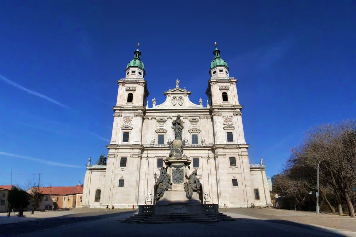 White Baroque cathedral with twin towers and green domes against clear blue sky, with monument in foreground in European city
