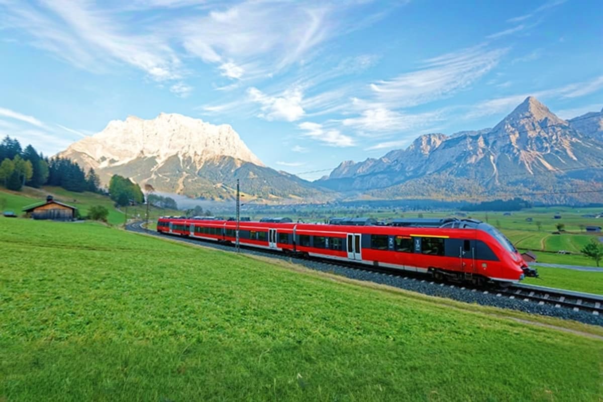 Red passenger train traveling through Alpine meadow with snow-capped mountains and lake in background, Bavaria region