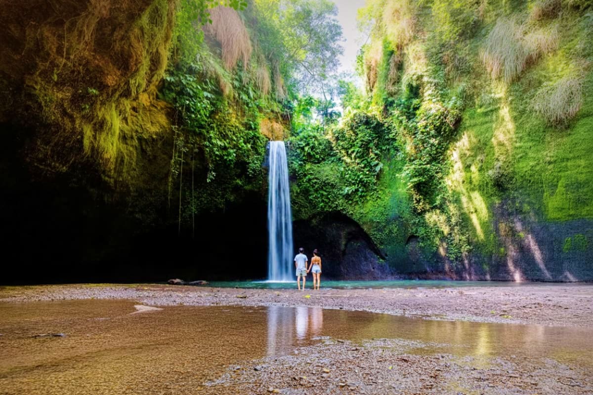 Two tourists standing on rocky beach viewing tall waterfall cascading from moss-covered cliff canyon with lush green vegetati