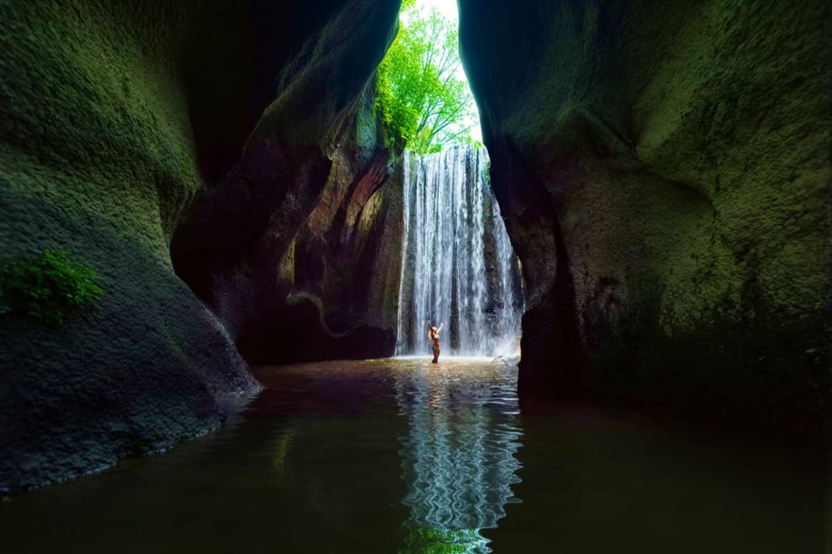 Person standing in shallow water beneath a waterfall cascading from a cave opening with lush green vegetation visible above