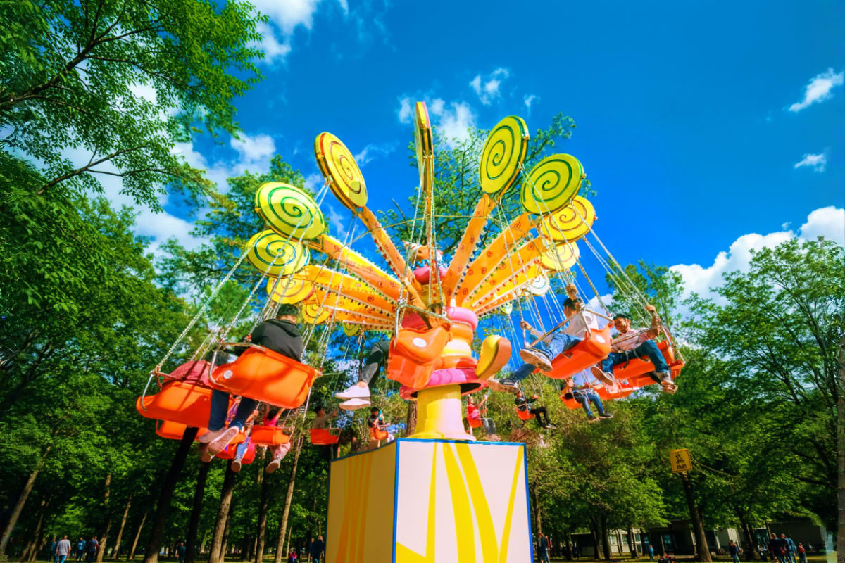 Colorful amusement park ride with bright yellow spiral seats and orange pods in motion against blue sky surrounded by green t