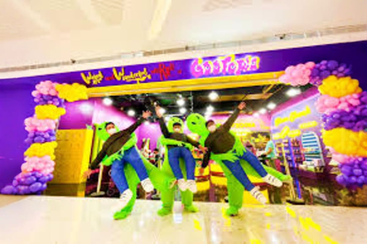 Three people in bright green costumes posing and dancing in front of colorful Gootopia entrance with purple and yellow decora