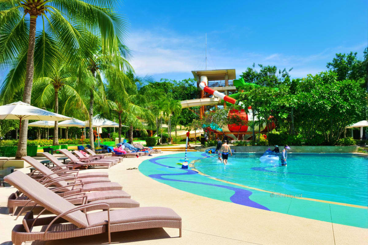 Tropical resort waterpark with turquoise pool, colorful water slide tower, lounge chairs, palm trees, and families swimming u
